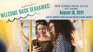 Two college women are riding the bus and looking down at a phone. Text readers Welcome Back Seahawks! The UNCW Seahawk Shuttle will begin August 18, 2021.